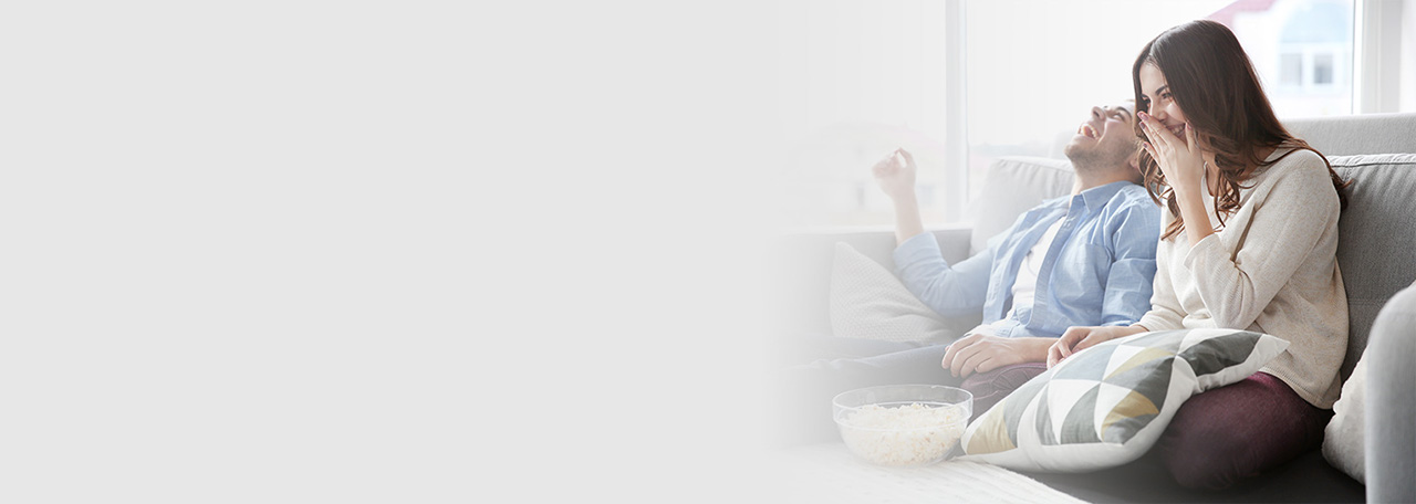 Пара сидит на диване, смеется и держит попкорн.